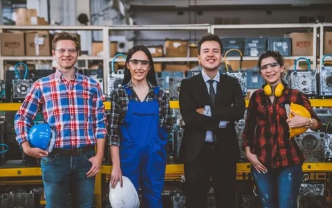 Four smiling factory employees in work clothes, safety glasses and a suit standing in a parts warehouse