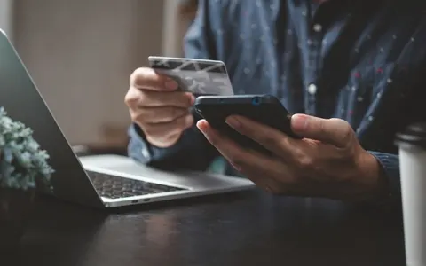 A person holding a bank card and a smartphone in front of a laptop, making an online payment