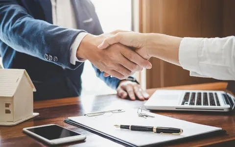 An adviser and a client shaking hands over a contract, a laptop and a small model house