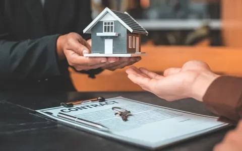 An agent handing a small model house to a client over a signed contract and keys