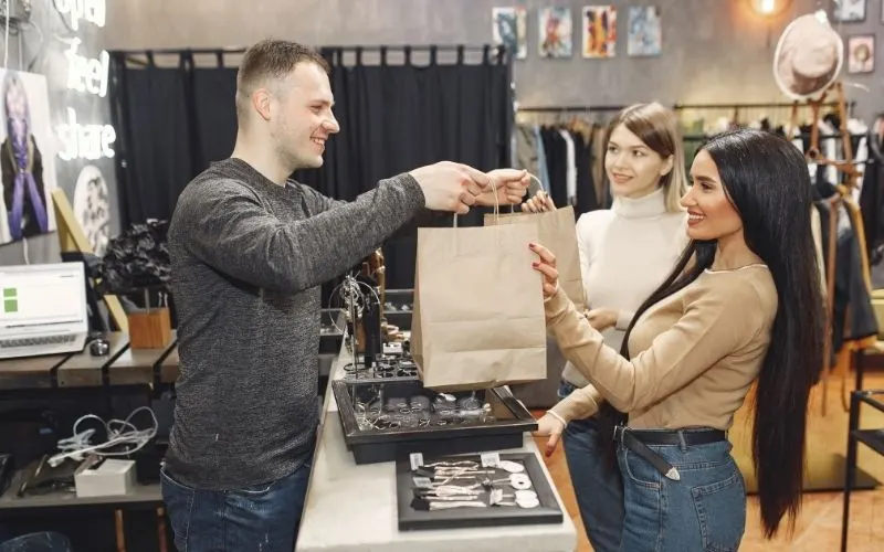 A sales assistant handing a shopping bag to two smiling customers in a clothing store